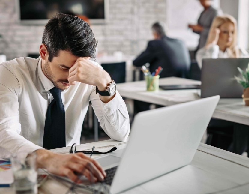 Tired businessman holding his head in pain while working on laptop and feeling exhausted from work. There are people in the background.